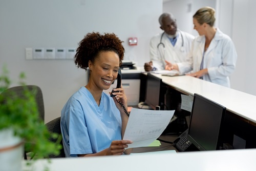 Mixed race female doctor at reception desk in a hospital holding a document and talking on the phone.