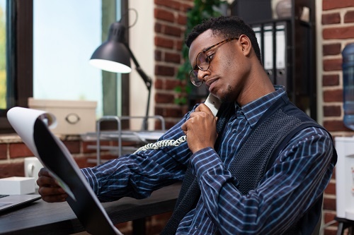 A black man in an office looking at a chart with a phone to his ear. 