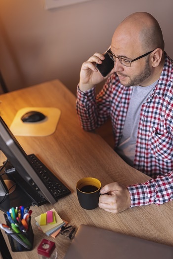 High angle view of a man speaking on the phone and drinking coffee in front of a computer screen.