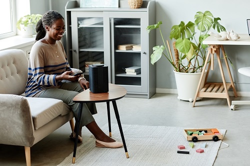 Portrait of smiling African American woman using voice activated smart speaker in cozy home interior.